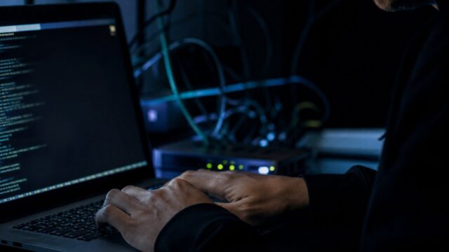 Hands typing on a laptop in a dark room lit by a terminal window